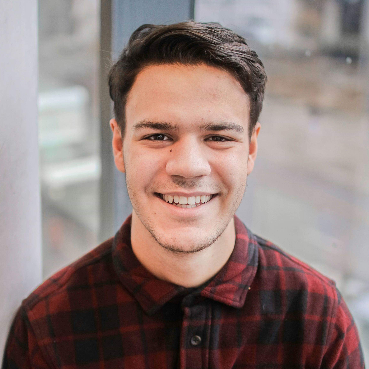 Nick, a young woman with short black hair, wearing a red plaid button-up shirt. He is looking into the camera and smiling.