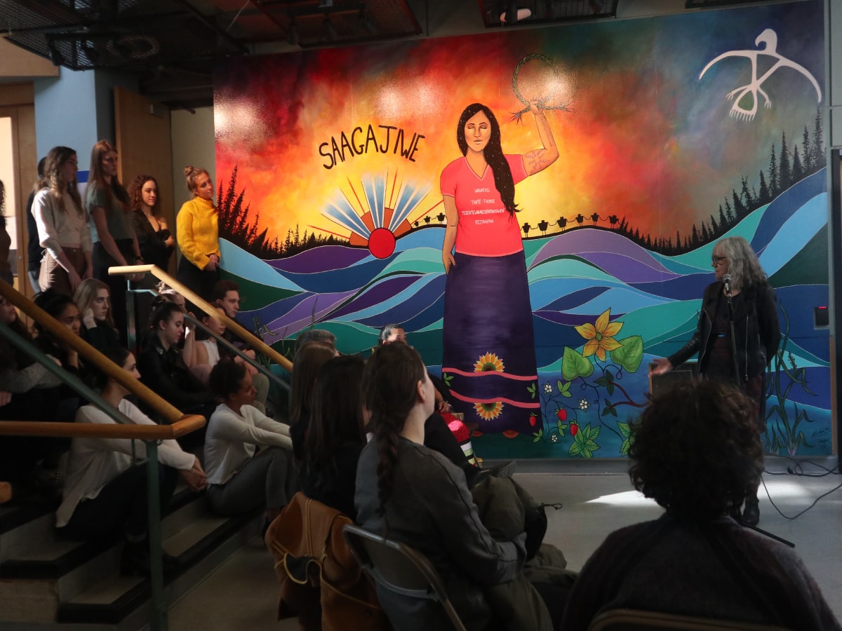 A mural featuring Indigenous art with a woman in the middle and a hill landscape behind her. On the left, the Saagajiwe logo is painted 