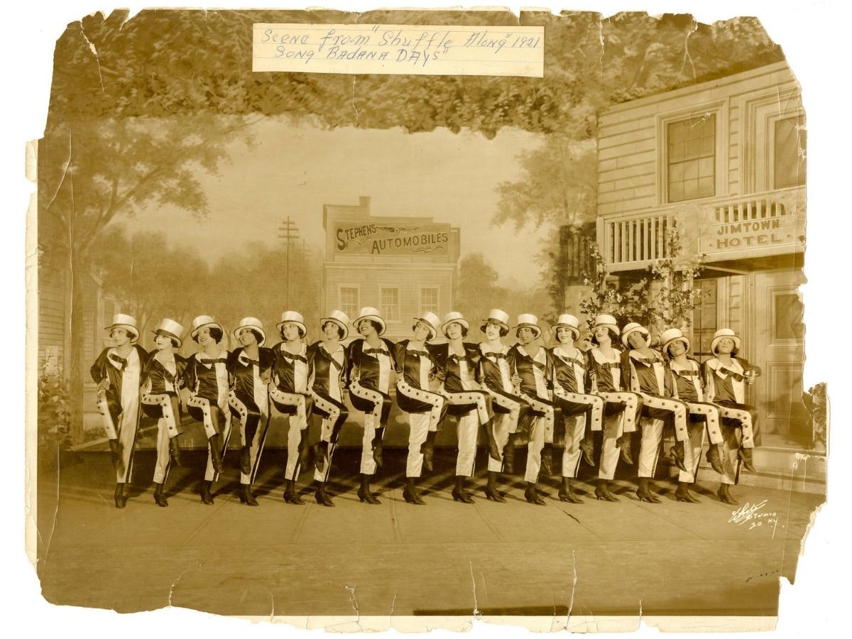 line-up of women on stage, standing side to side with their arms draped around eachothers shoulders and one leg off the ground