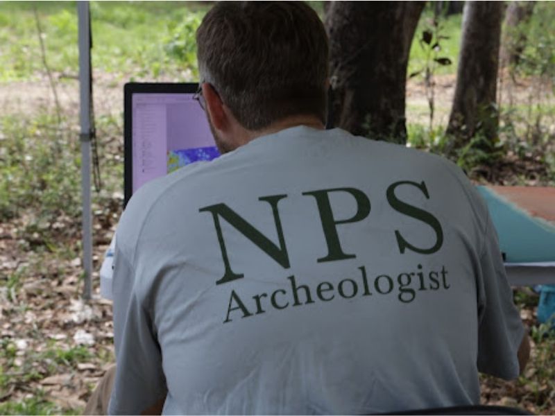 Man sitting outside, facing away wearing a grey shirt and looking at a computer screen