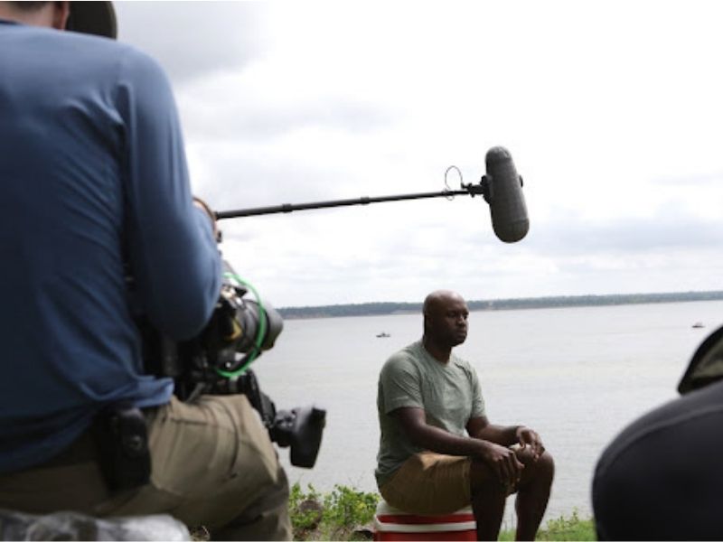 The back of a man wearing a blue shirt pointing a camera at a man sitting on a red cooler wearing a grey shirt and khaki shorts, staring off screen with a boom mic over his head