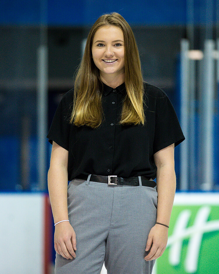 Camryn, a young woman with long brown hair, wearing a black button-up shirt and grey trousers. She is looking into the camera and smiling.