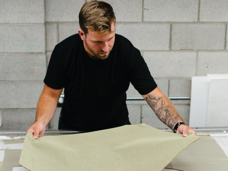 Fashion alum Ben Christy is wearing a black t shirt and he is looking down at a piece of cut beige fabric. He has brown hair and a half sleeve of tattoos