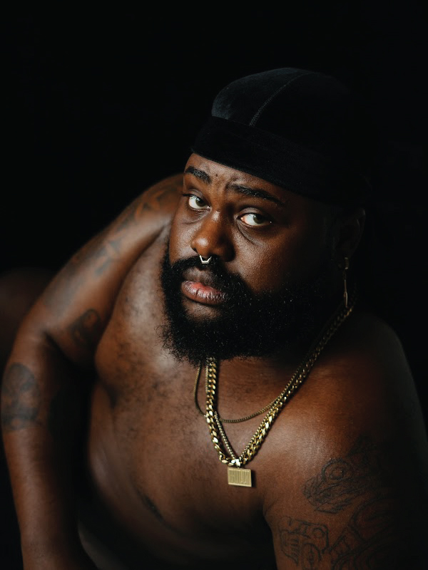 The photograph shows a shirtless black man with tattoos wearing gold chains, a septum ring, and a black durag on his head. He is posed lying on his side, perched up on his elbow in front of a black background. He looks directly at the camera with a neutral face.