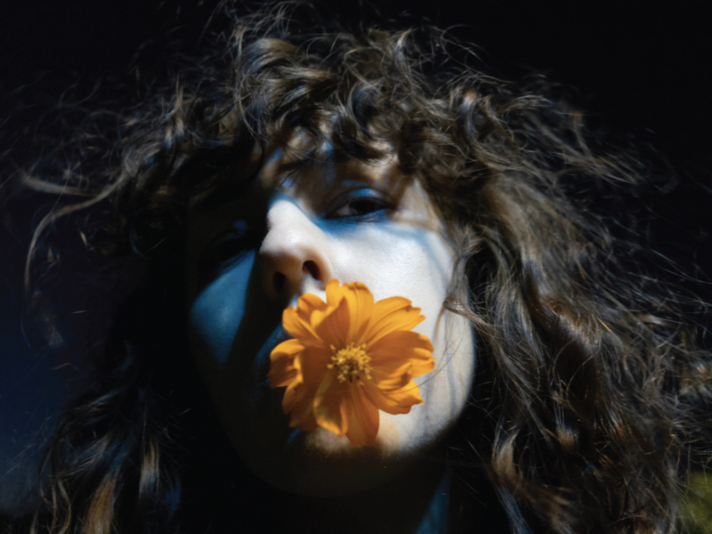 The photograph shows Ilina with an orange flower in her mouth, captured from a low angle. Her brown curly hair is illuminated and a blue light casts a shadow over her eyes. 