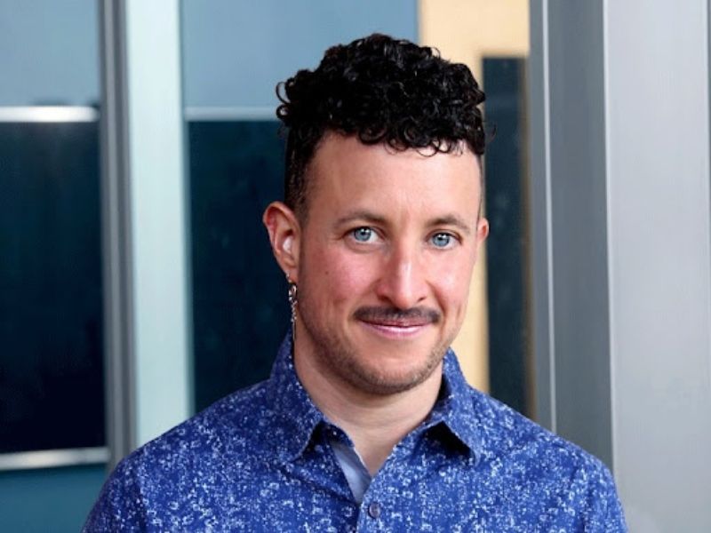 Portrait of Marty Fink. Fink wears a blue button down collared shirt and smiles with their mouth closed for the camera. They have a silver drop earring in one ear and have short black curly hair.
