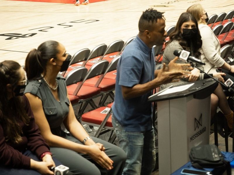 A man talks at a podium addressing students in the bleachers at an arena