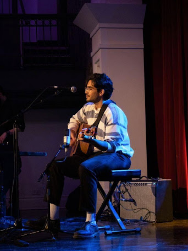 A man sitting on a chair playing guitar and sitting to an audience that is off the screen.