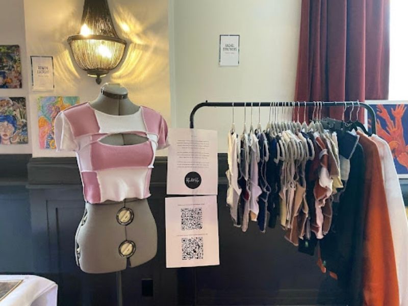 A mannequin adorning a white and pink crop top. To the right is a clothing rack with colourful clothing.
