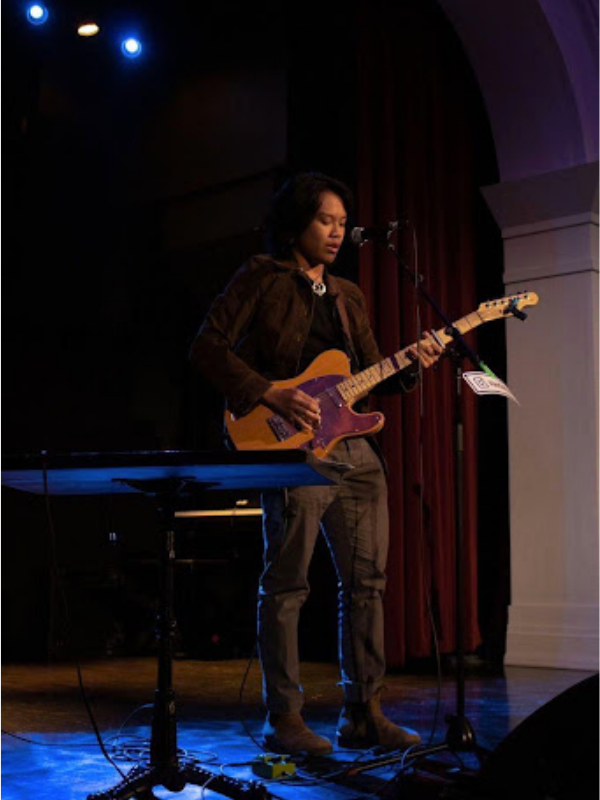 A man standing on a stage holding an electric guitar and singing to an audience of people.