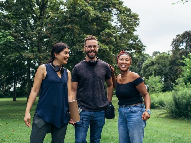 Three people standing outside smiling and staring into the camera. The background is full of lush greenery.