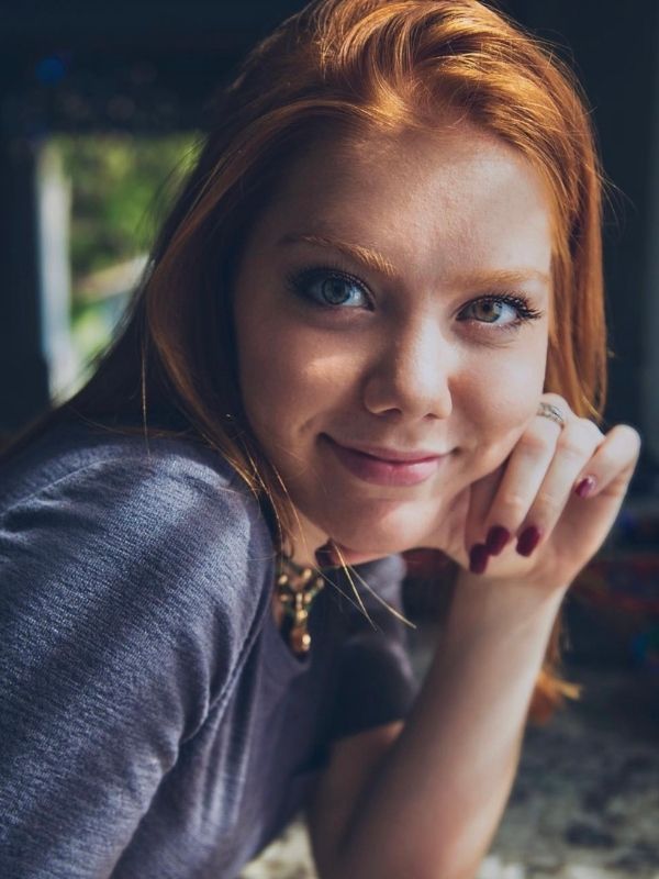 Young woman's headshot, She wears a grey textured shirt and gold necklace, and has one hand against her cheek. Her nails are painted red.