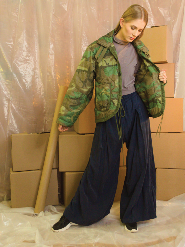 A woman wears baggy black pants and an army green puffer jacket. She stands in front of a stack of boxes