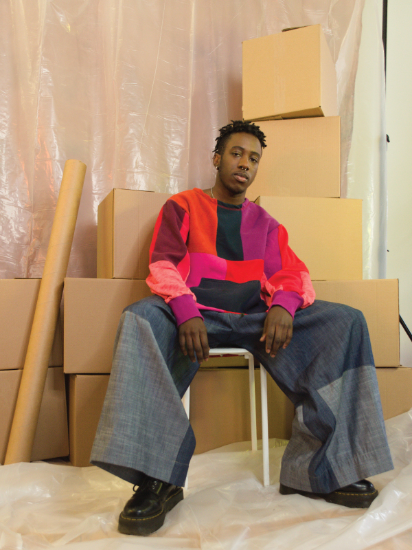 A man wears a red and pink block patterened sweater. He sits in front of a stack of cardboard boxes