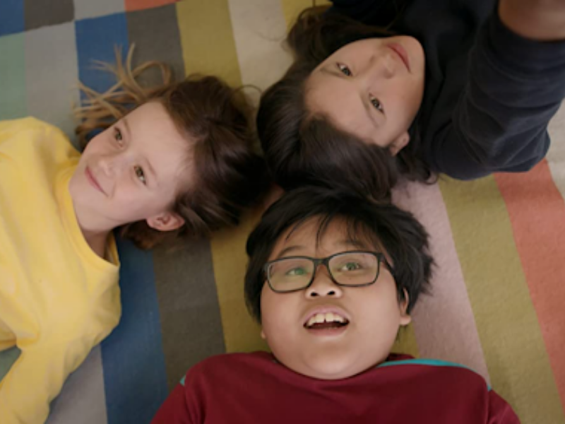 Still from a movie depicting three children laying on their back staring off into the distance. The three kids are laying on a brightly coloured tapestry.
