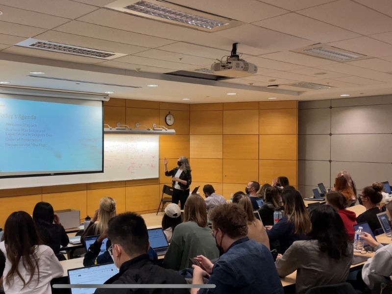 Students in a lecture hall watch a presentation being made by a woman who is pointing at slides on a screen in front of the class