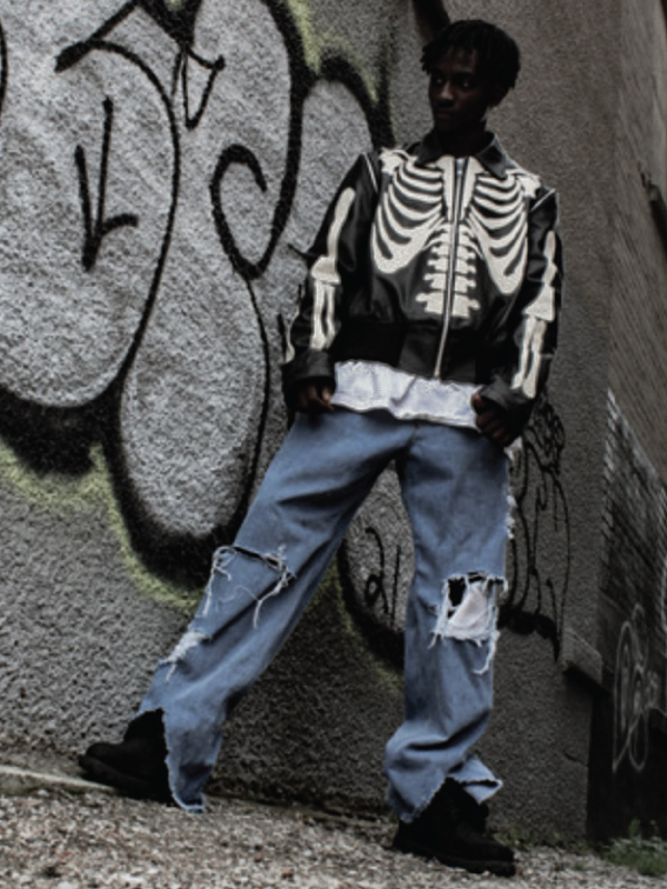 A man stands in front of a grafitti wall wearing blue jeans and a black skeleton jacket