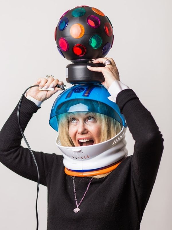 Blonde woman wearing a plastic helmet attempting to plug in a colourful stroblight with one hand while holding th elight on top of her head with the other