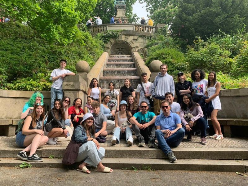 : A group of 20 to 25 students and instructors are posing on stairs in front of a large water fountain. 