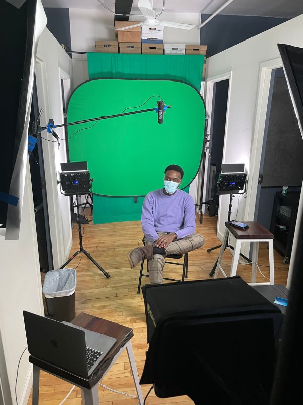 A filming room with one man sitting down in a chair wearing a face fasm. Behind him is a green screen and in front there is a teleprompter.