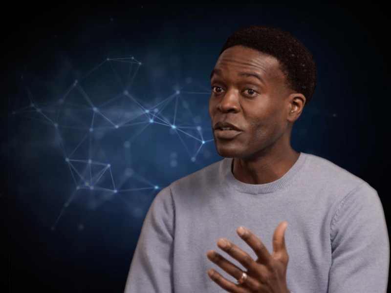 A man wearing a grey sweater holds his hand up expressively. He sits in front of a dar blue background.