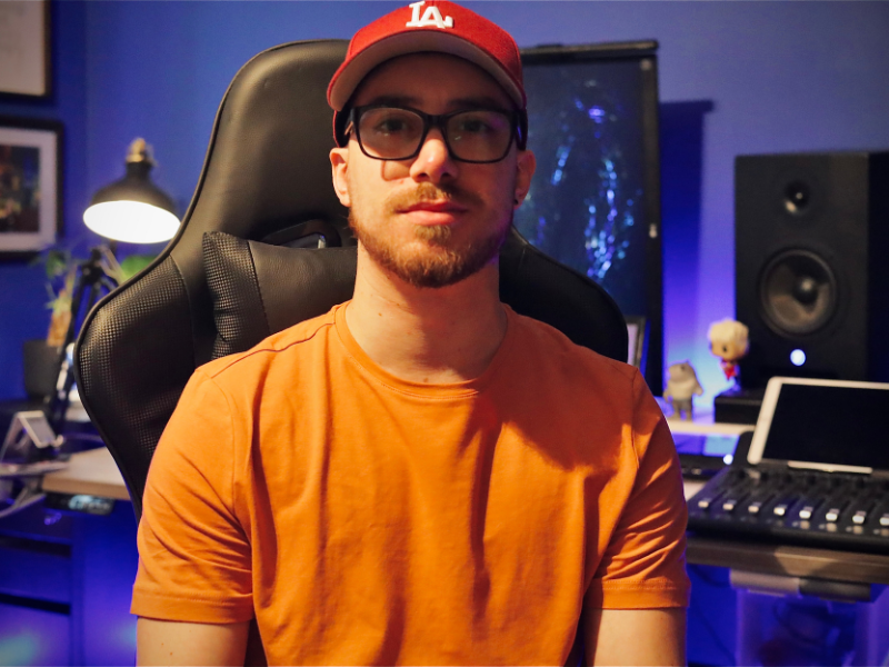 A man wearing glasses, an orange shirt, and a red baseball cap is sitting in a chair surrounded by audio equipment. 