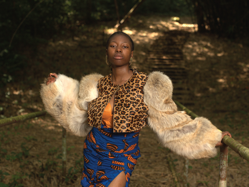 An African woman poses in a forest wearing a cheetah printed top, fur coat and blue and oorange printed skirt