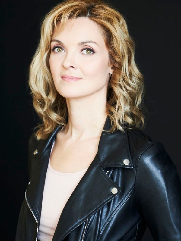 A woman with blonde hair and wearing a black leather jacket faces the camera. Her Jacket partially blends into the black background.