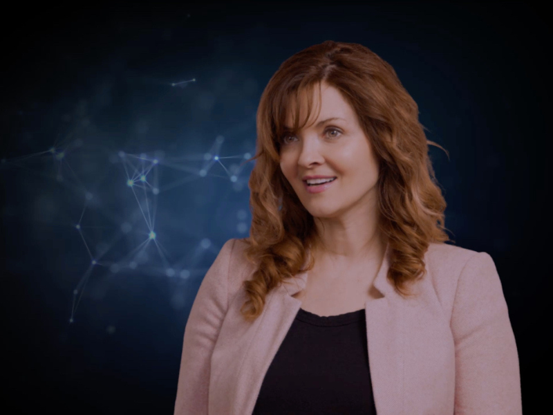 A woman smiles and looks off into a camera. She is wearing a pink blazer which sharply contrasts with the dark blue background.