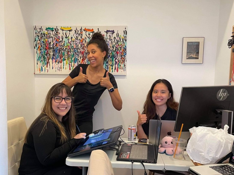 Three women are gathered around a desk with laptops in front of them holding their thumbs up 