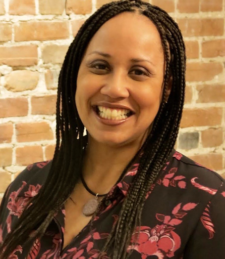 A woman wearing a floral blouse stands in front of a brick wall and smiles.
