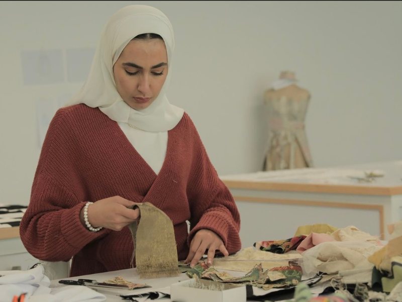 A woman in a hijab is standing behind a desk covered in fabric, holding up a piece. 
