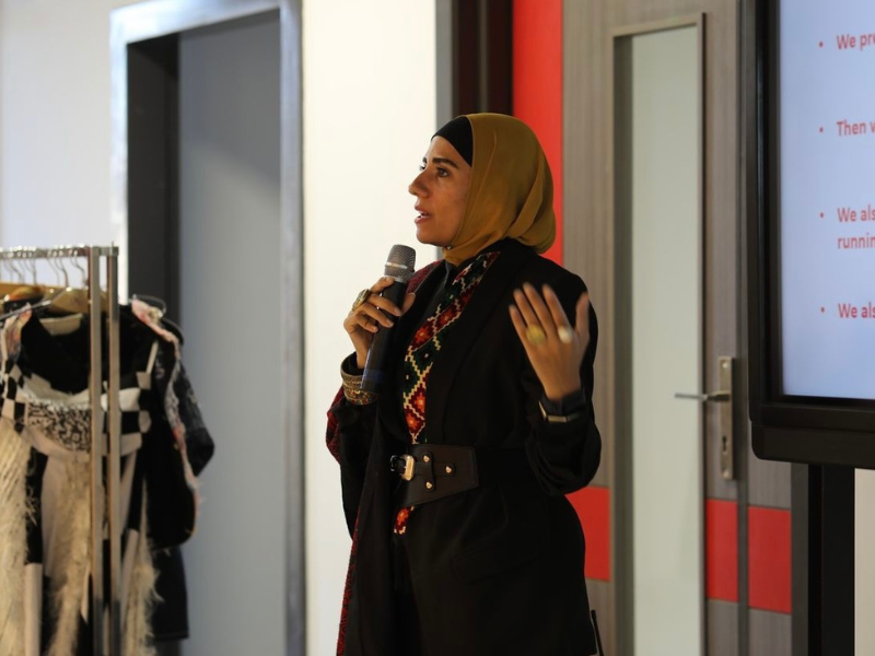 A woman in a hijab is holding a microphone and presenting infornt of a slide deck 