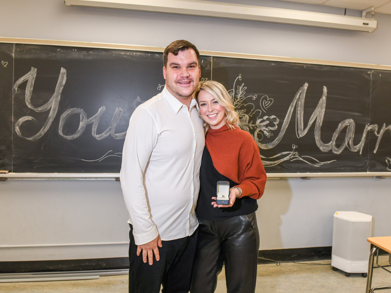 A man is hugging a woman's side. The woman has a an open ring box in her hand 