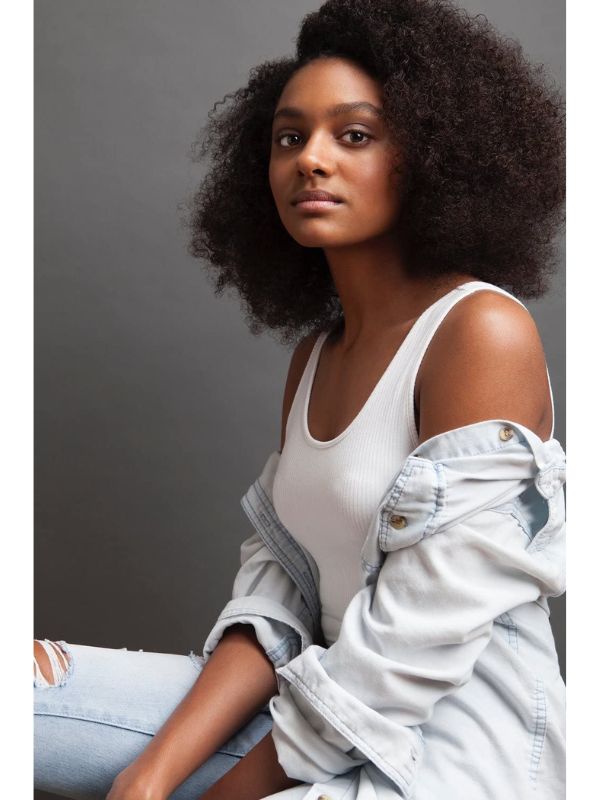 A woman sits up straight and stares into the camera. She is wearing a white shirt with a gray sweater draped over her arm.