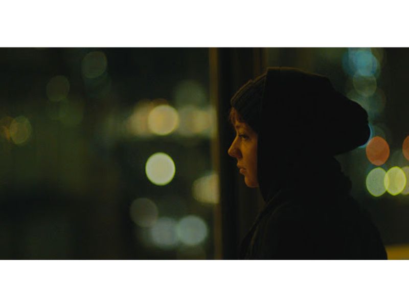 A woman wearing a hoodie stares into the distance with bokeh lights glistening the background.