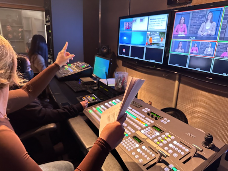 A woman sitting in a production room holding a sheet of papers in front of two television screens.