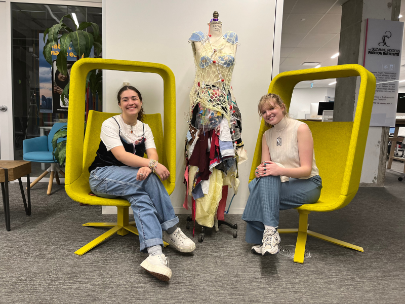 Two students sit beside a mannequin adorned in a dress made of upcycled medical waste