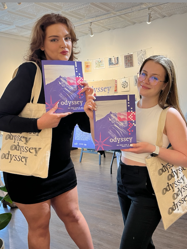 Two students are holding up posters of the Odyssey exhibit in a gallery space and wearing Odyssey-branded totes.
