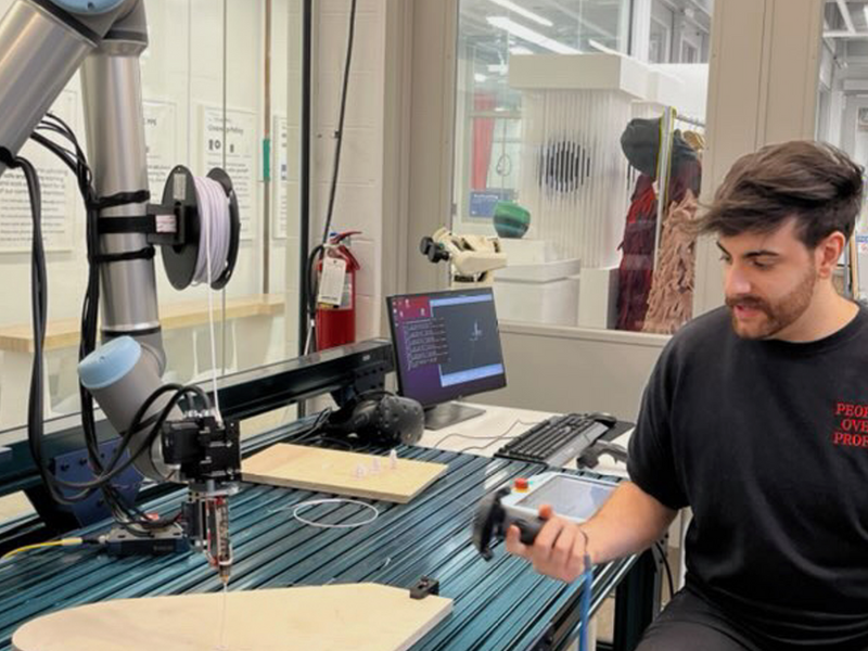 A student is holding a controller in his hand to maneuver the robotic arm next to him, directing it to 3D print.