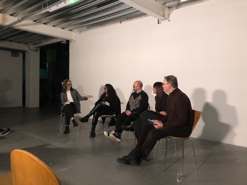 Natalie Alvarez, Associate Dean, Scholarly, Research, and Creative Activities, at The Creative School, is sitting down with the artists involved in the exhibit on her right. There is a mic in her hand as she moderates the discussion.