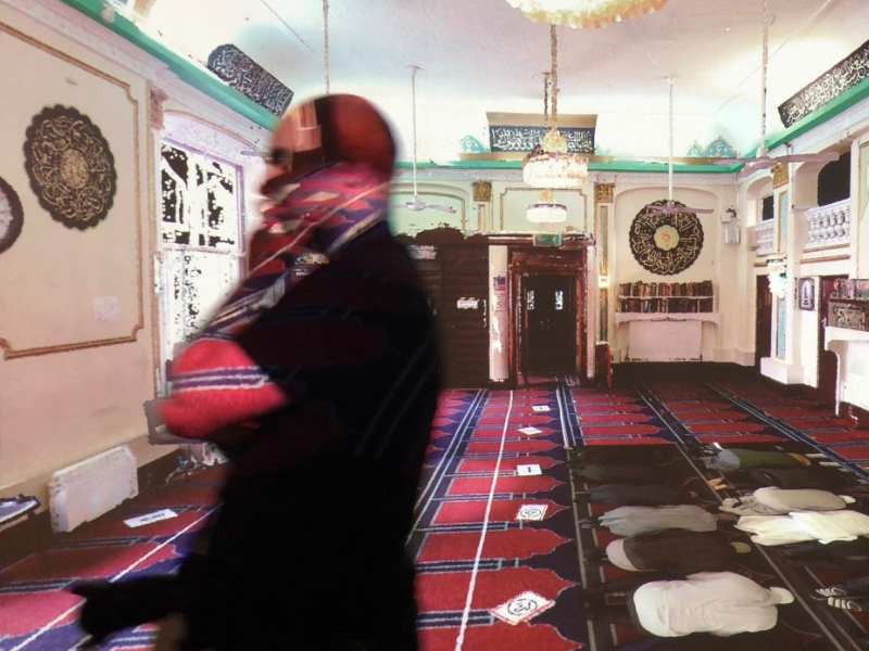 A person is walking in the foreground inside a mosque. Behind them, people are lined up praying.