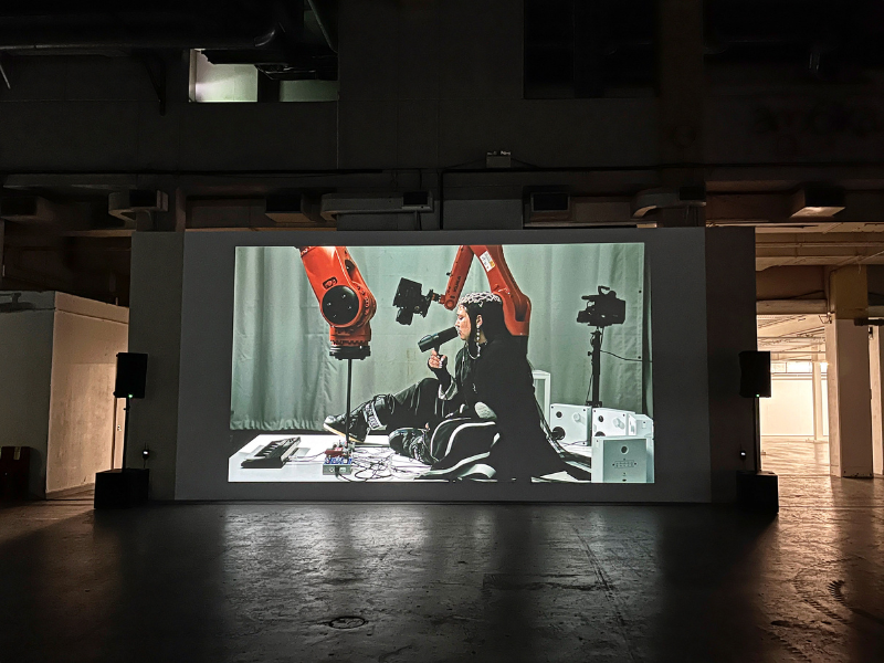 A projected image on a dark wall of a woman sitting next to a large orange robotic arm. The woman is holding up a microphone to her mouth, the large robotic arm is pressing a key on a piano keyboard.