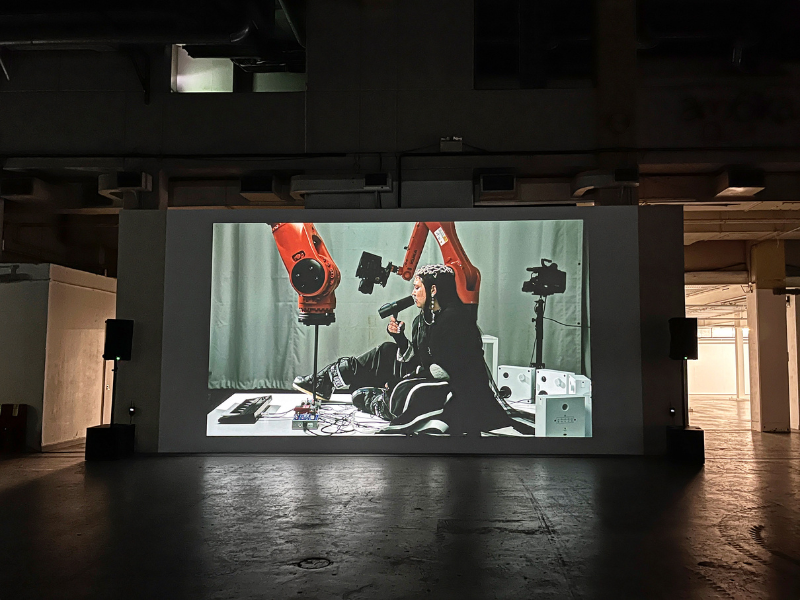 A projected image on a dark wall of a woman sitting next to a large orange robotic arm. The woman is holding up a microphone to her mouth, the large robotic arm is pressing a key on a piano keyboard. 