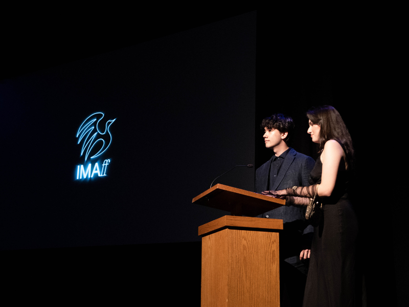 Two students, Katie wearing a dress and Gira wearing a suit, are standing in front of a wood podium. The word IMAFF is projected behind them on a screen.  