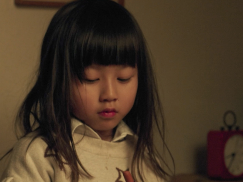 A still image of a little girl with black hair and bangs dressed shirt and vest looking down. 