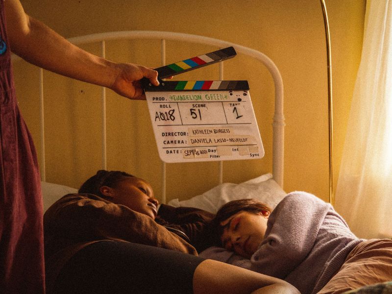 Behind the scenes of a film shot with two actors lying in a bed and a clapper signifying the beginning of a take.