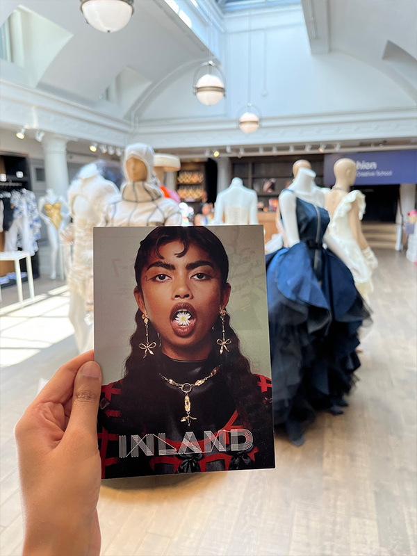 A hand is holding up a postcard. Printed on the card is an open-mouthed woman with a flower on her tongue. INLAND is written at the bottom of the card. 