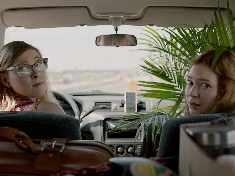Two actors sit in a car and look behind through their back window
