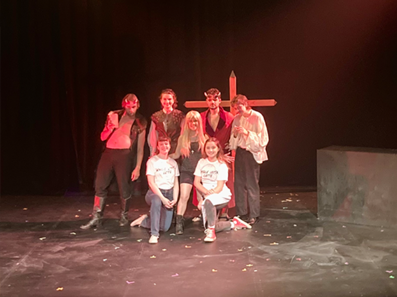 The cast of the show is posing on the stage with a large cross behind them